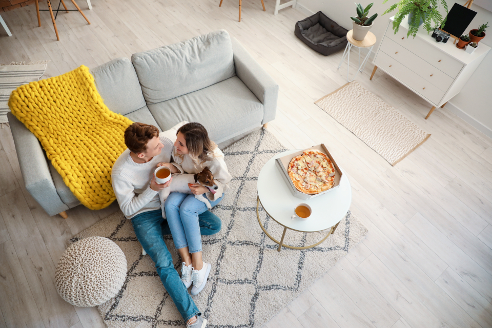 Couple sitting by a couch with pizza and warm drinks for an at-home date night.